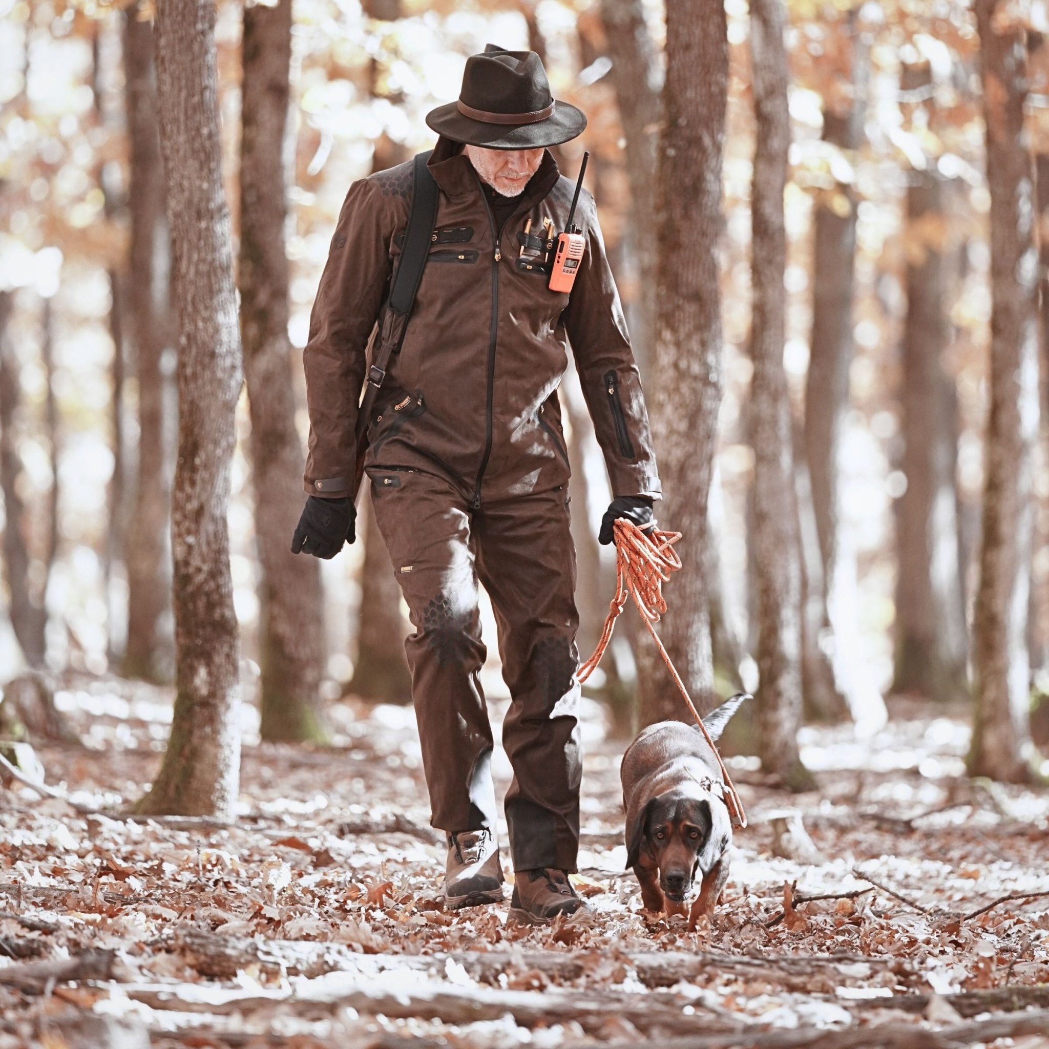 A person in a brown outfit and hat, wearing HILLMAN GEARs 6OL Matrix Insulated Hunting Trousers | Waterproof Bibs, walks a dog on a leash through the forest. Fallen leaves cover the ground as sparse trees surround them. They carry a device with an antenna over their shoulder.