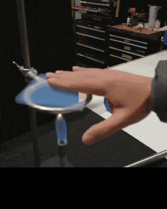 A person holding a blue object with a circular rim and a white bottom, balancing it on a fingertip. The background shows a black table and shelves with tools.