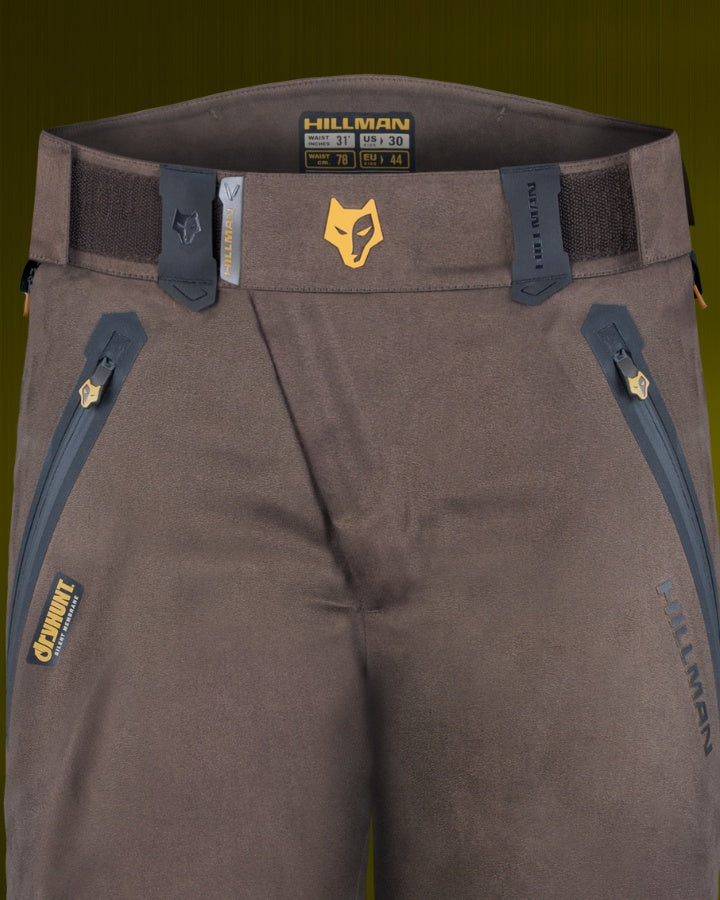 Close-up of brown Hillman outdoor pants showcasing the brands logo and zipped pockets on a green background. The waist size is indicated on the label, and adjustable straps are visible at the waistband.