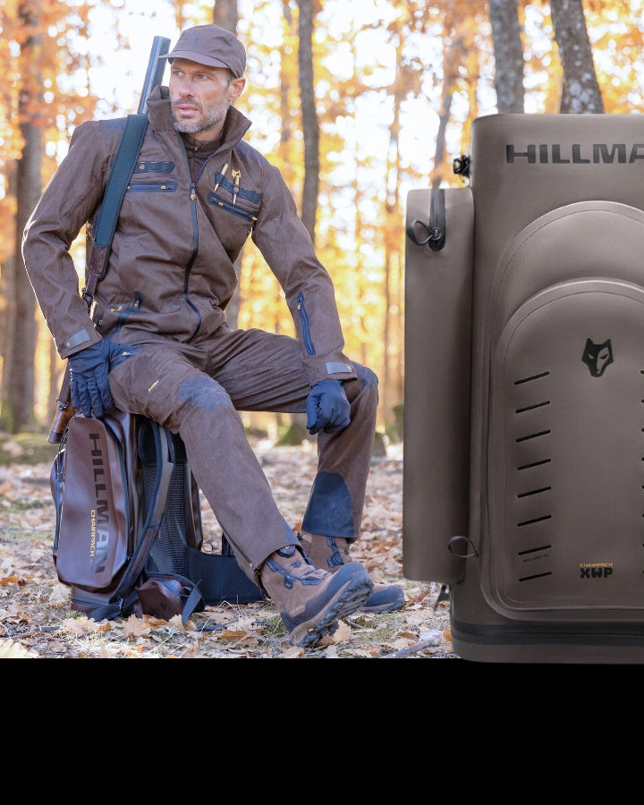 A person in brown outdoor gear crouches in a wooded area with fallen leaves. They hold a rifle and have a backpack beside them. The scene includes a close-up of a backpack with HILLMAN printed on it. The setting appears to be autumn.