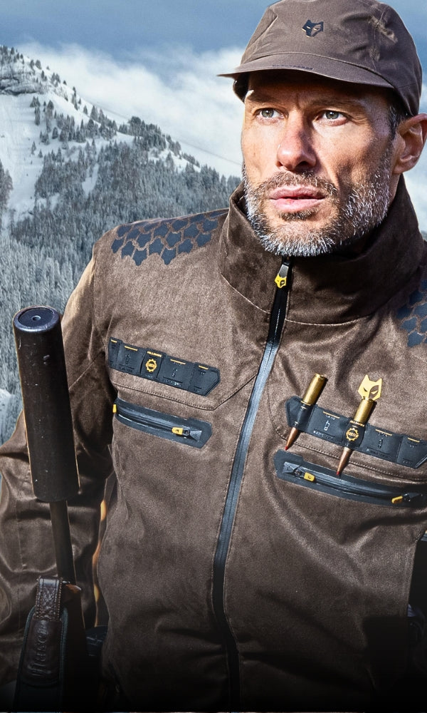 A man wearing a brown jacket and cap stands with a snowy mountain in the background. The jacket features multiple pockets and a zipper. The sky is cloudy, hinting at cold weather.