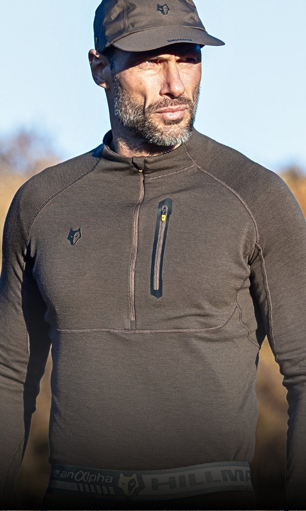 A man in a green long-sleeve, zippered athletic top and matching cap stands outdoors. The top has a small chest pocket. The background is a blurred natural landscape under clear blue skies.