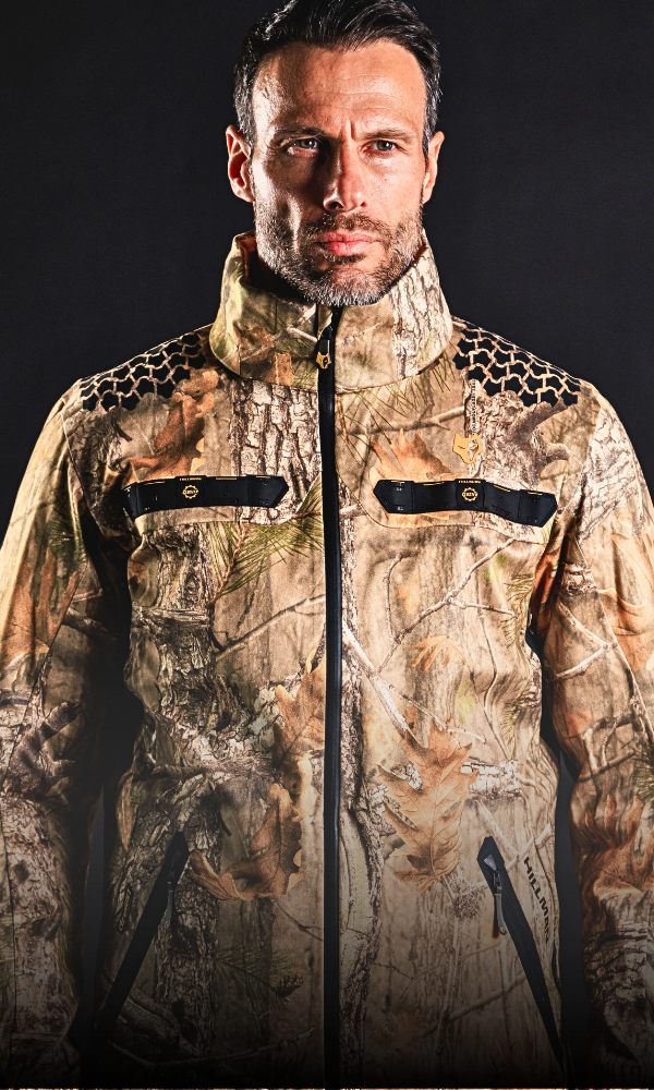A person wearing a camouflage jacket with dark zippers and a high collar, standing against a solid black background.