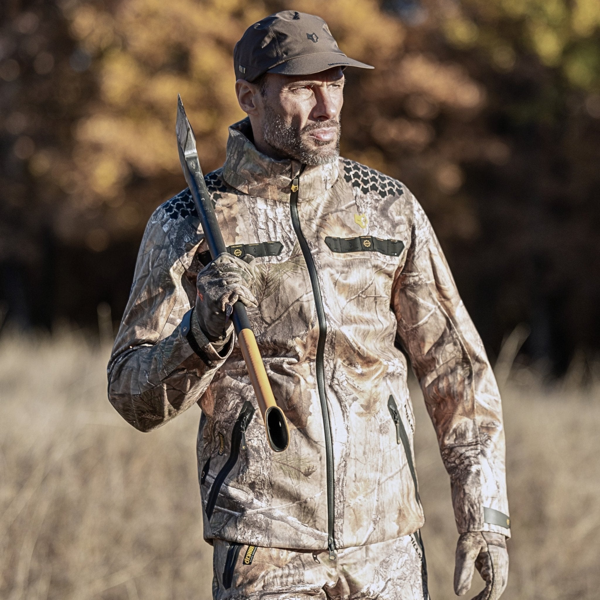 A man in camouflage hunting gear and cap stands outdoors holding a spear, wearing the HILLMAN GEAR ultra-lightweight camo rain jacket | Waterproof Camouflage Hunting Coat. Dry grass and blurred autumn trees are visible in the background.