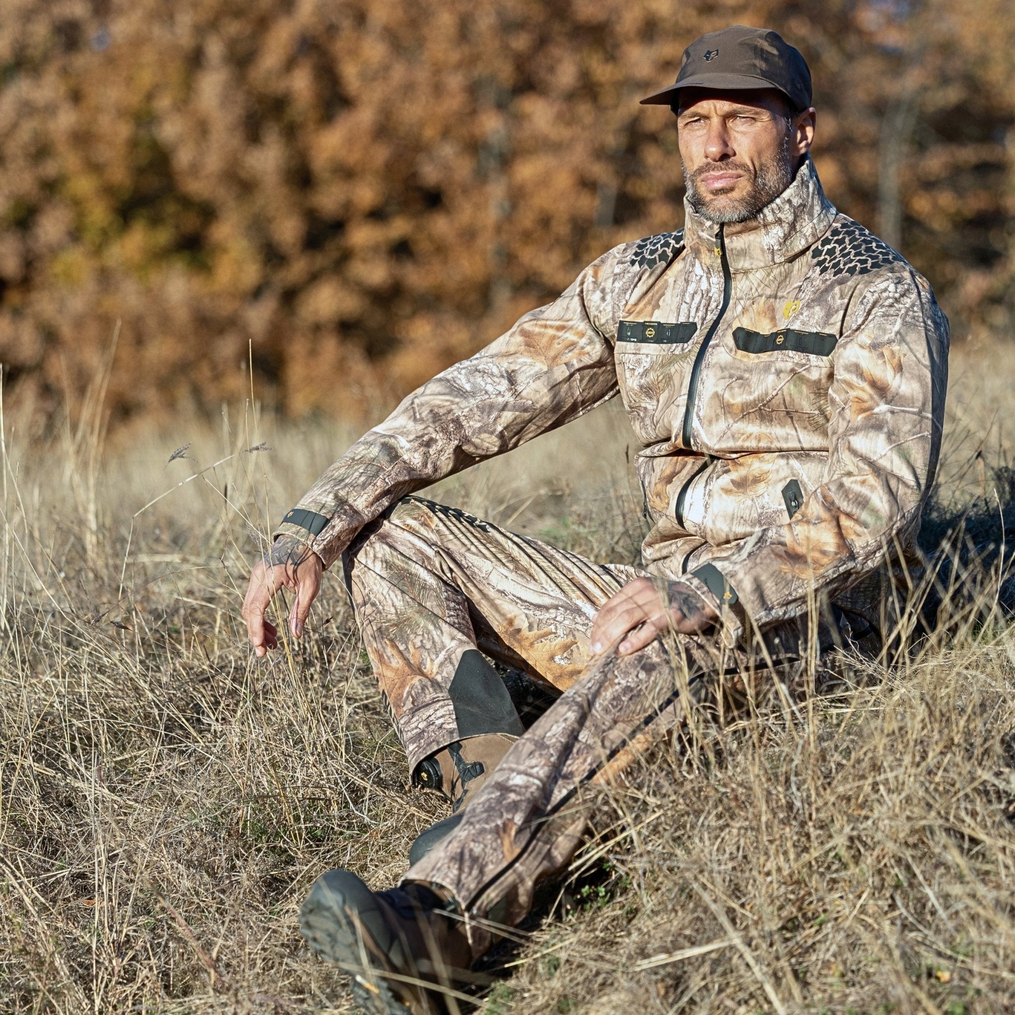 A man wearing a HILLMAN GEAR ultra-lightweight camo rain jacket and cap sits on dry grass in a field with brown autumn trees in the background, looking slightly off to the side.