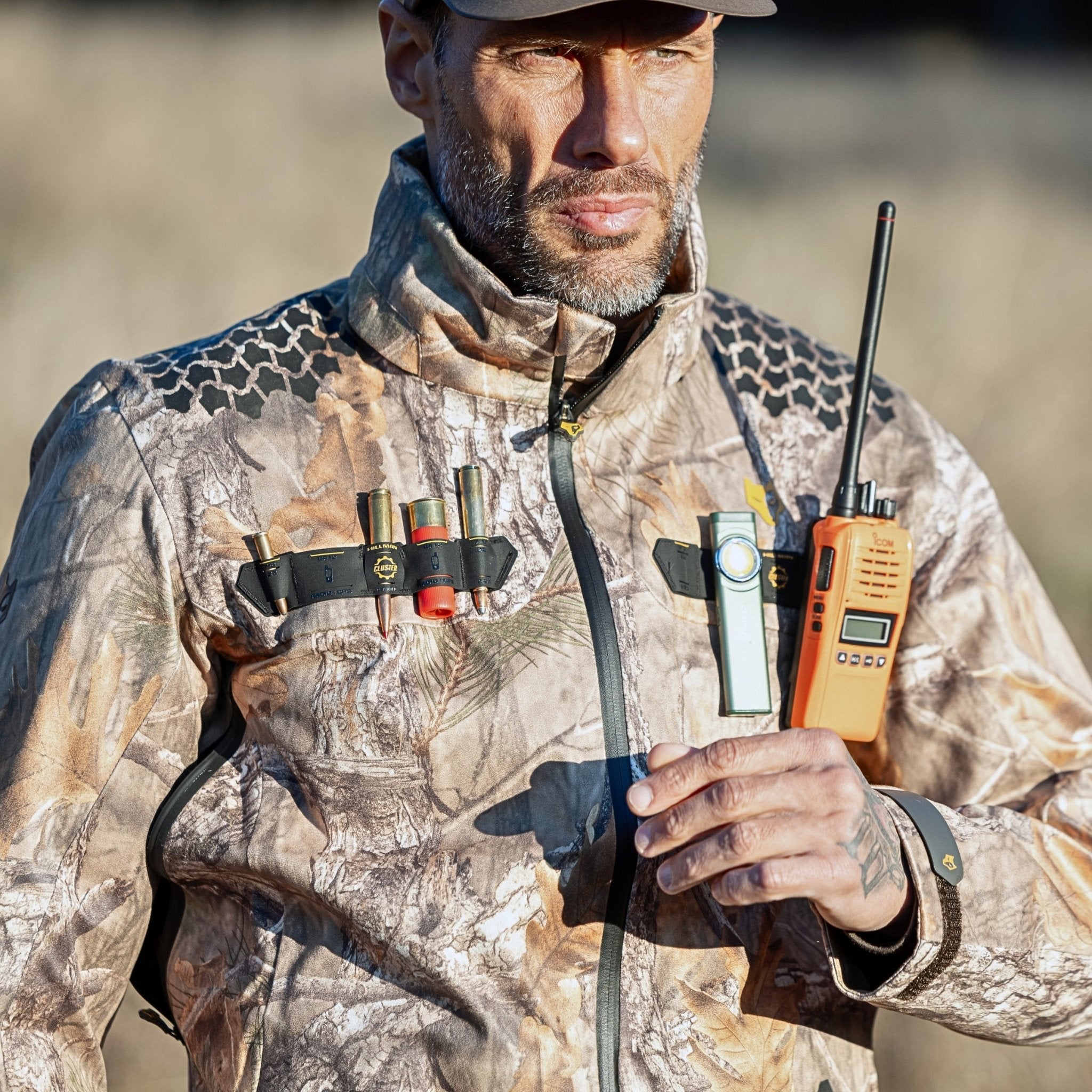 A man wears the HILLMAN GEAR ultra-lightweight camo rain jacket, equipped with shotgun shells, a flashlight, and a whistle on the chest. He holds an orange walkie-talkie. The outdoor background is slightly blurred.