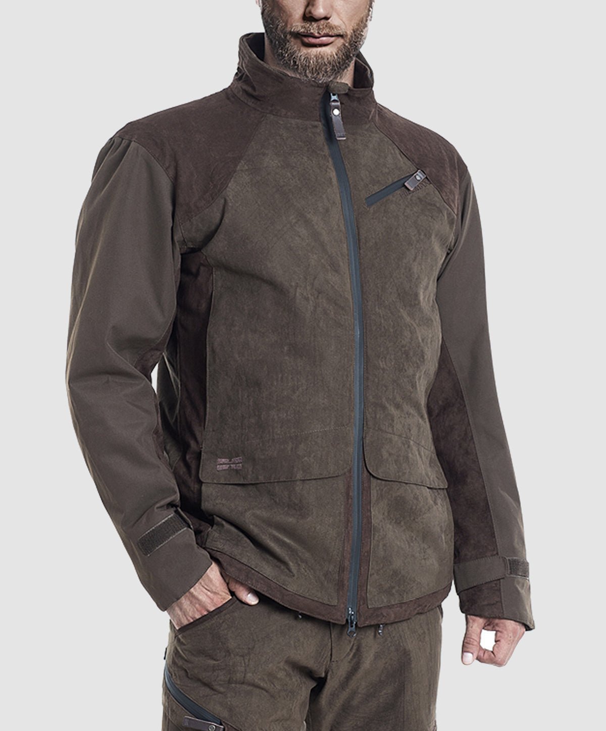 A man wears the Hillman® PRE ORDER Fusion Hunting Jacket—a brown, waterproof outdoor jacket with a high collar and zippered chest pocket—paired with matching brown pants, standing against a plain light gray background.