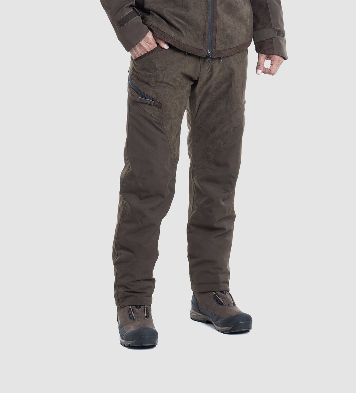 A person stands against a plain light gray background, wearing Hillman® PRE ORDER Fusion Waterproof Hunting Trousers paired with a brown outdoor jacket and dark hiking shoes.