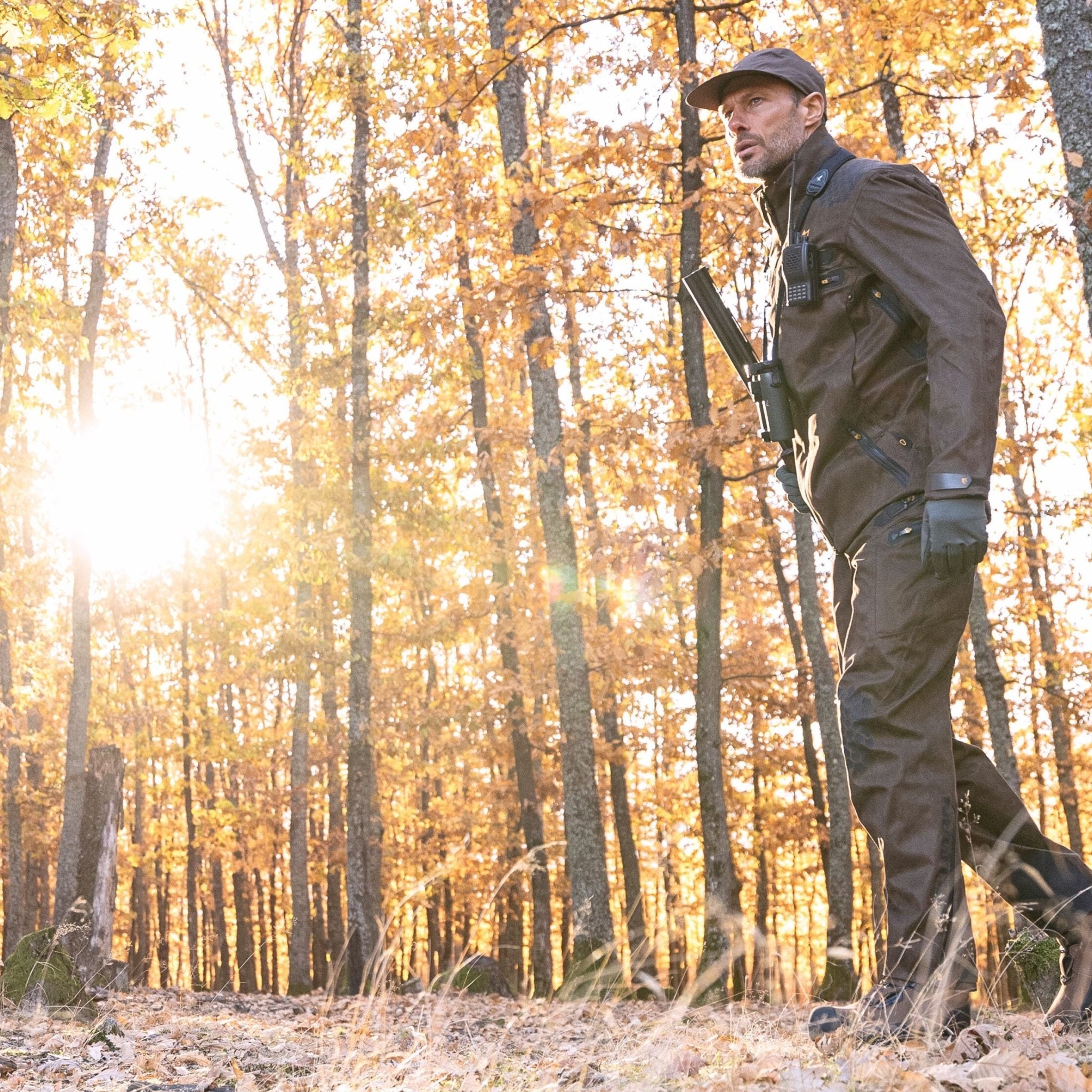 Wearing HILLMAN GEARs 6OL Matrix Insulated Hunting Trousers, a man in outdoor gear and a cap strolls through a sunlit forest with autumn leaves, carrying a long object over his shoulder as sunlight creates warm, golden hues through the trees.