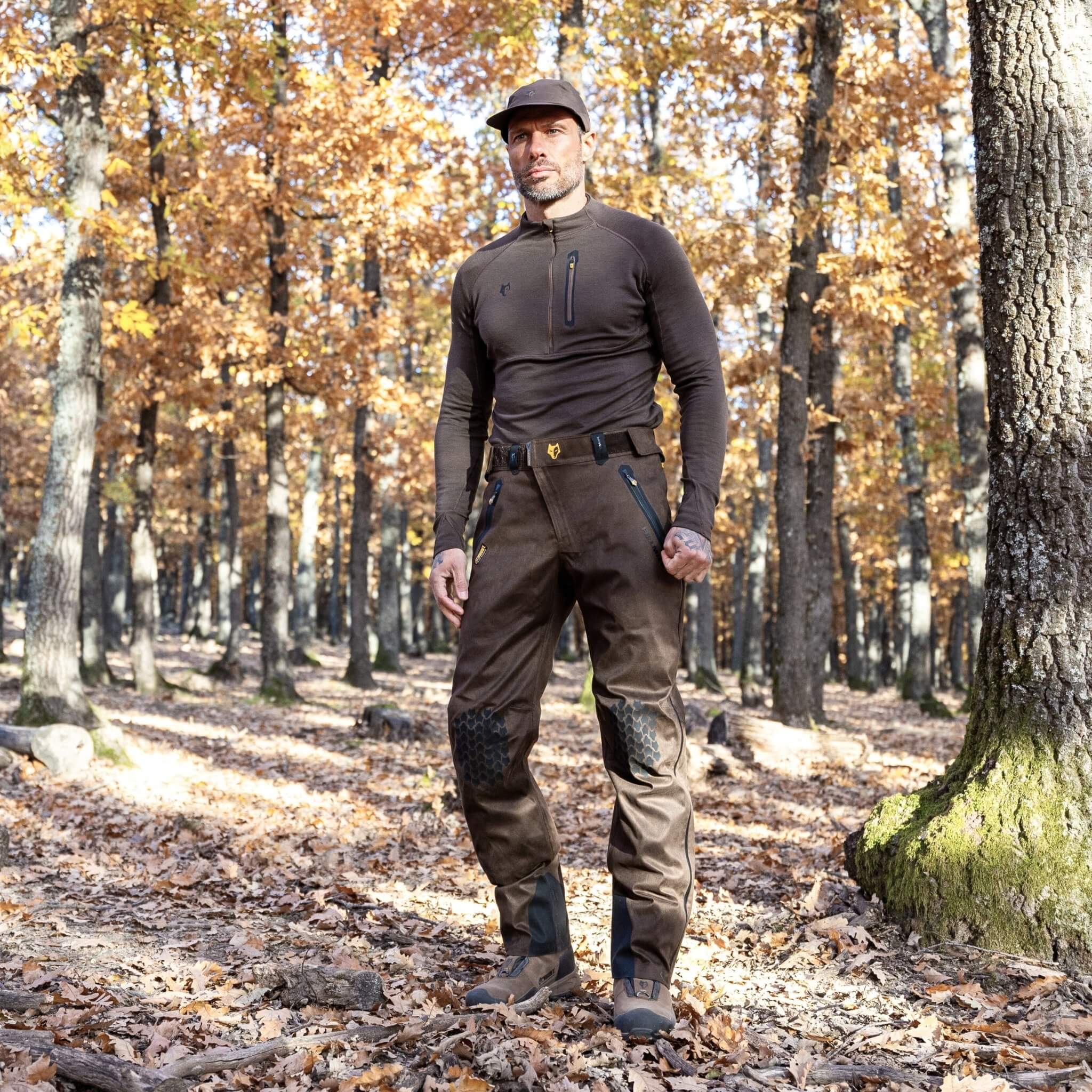 A person stands in a sunlit forest, surrounded by autumn leaves, wearing HILLMAN GEARs ultra-lightweight waterproof brown long-sleeve top, 5WL Ultra-Lightweight Waterproof Hunting Trousers with black knee patches, and a cap. The serene scene is enhanced by light filtering through the trees.