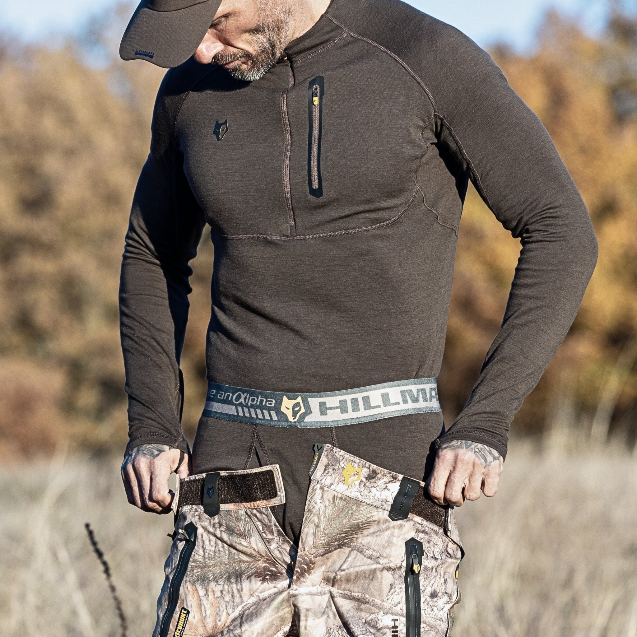 In a natural setting with trees and grass, a man adjusts his camouflage pants wearing a 2BL Merino Wool Hunting Long Johns thermal base layer shirt and cap. The waistband of the pants reads Hillman.