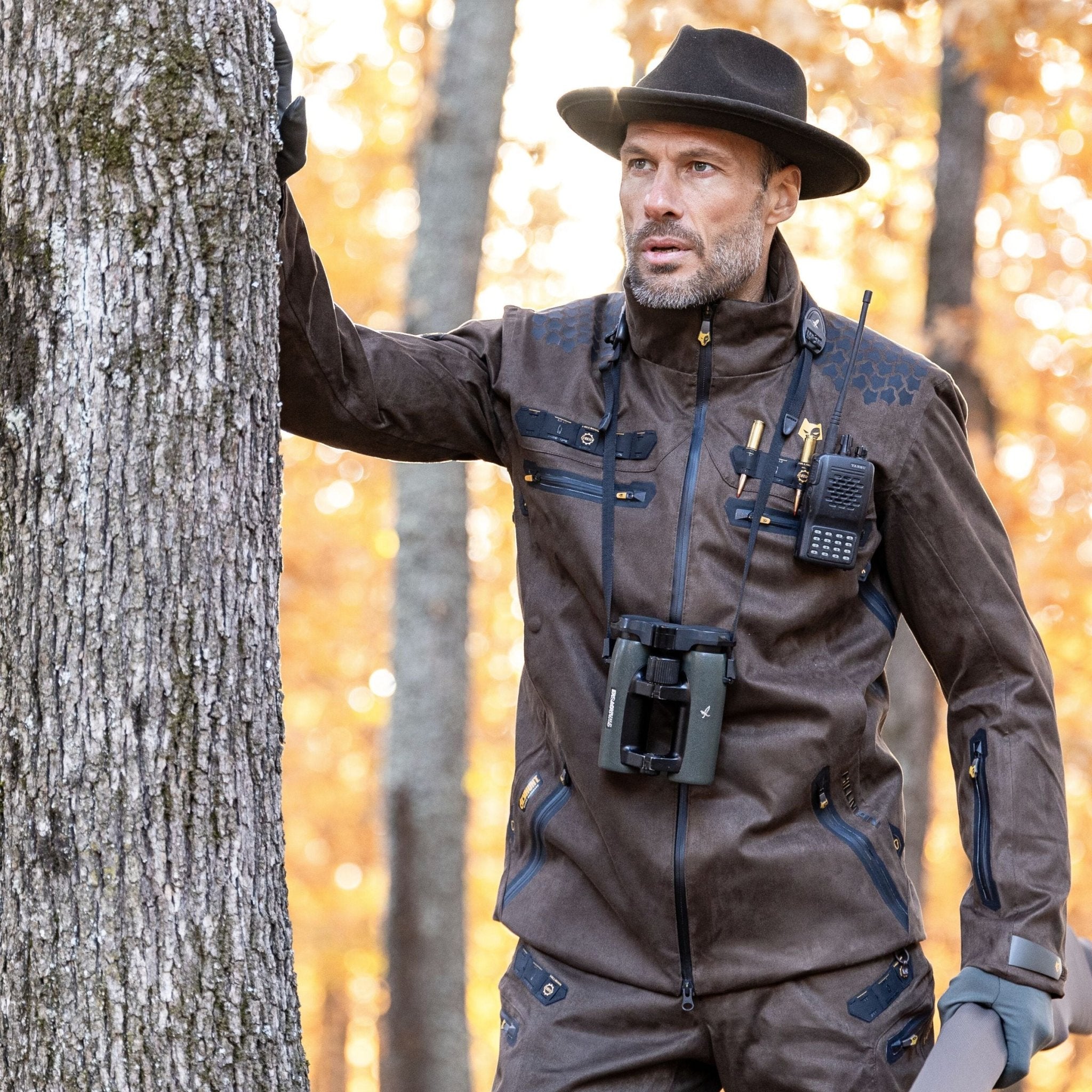A man in a brown HILLMAN GEAR heated hunting jacket, "The Warmest Heated Coat," and a black hat stands by a tree with autumn leaves, binoculars and walkie-talkie around his neck, looking into the distance.