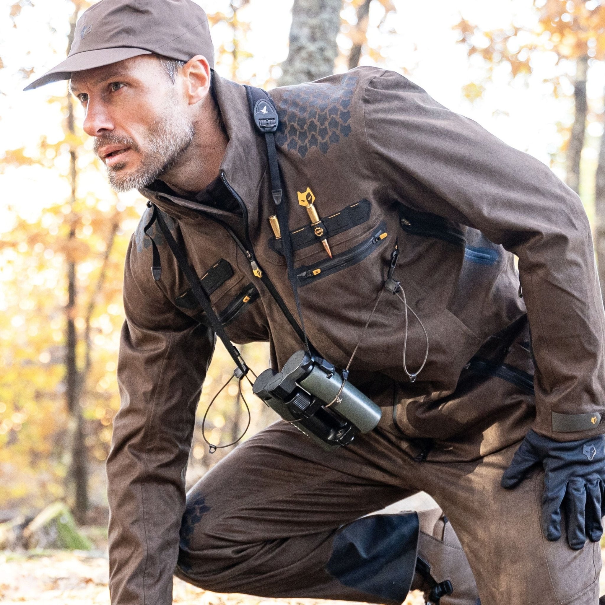 A man wearing the HILLMAN GEAR heated hunting jacket, The Warmest Heated Coat, kneels in a forest with binoculars around his neck, focused amid autumn trees and fallen leaves.