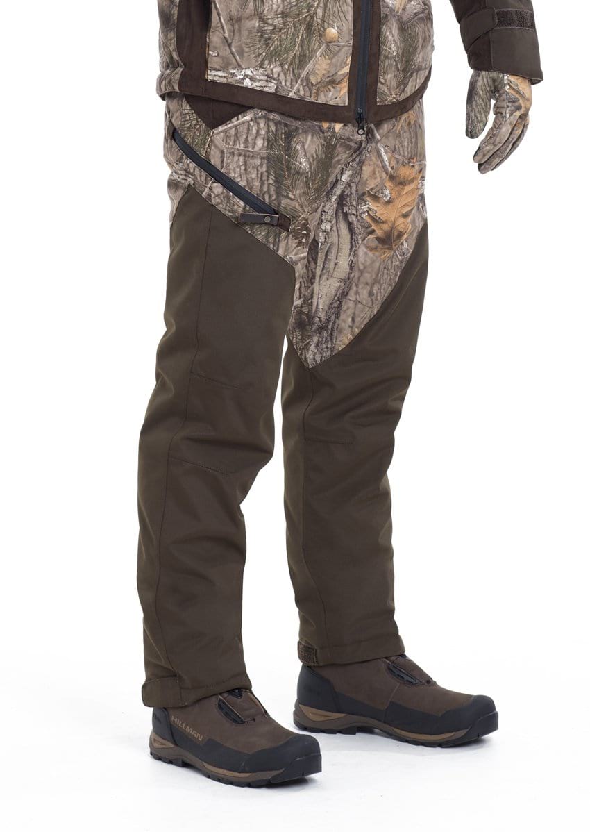 A person wears Hillman® fusion waterproof hunting trousers in camouflage and dark olive, paired with brown hiking shoes and camo gloves, standing against a white background with only the lower half of their body visible.