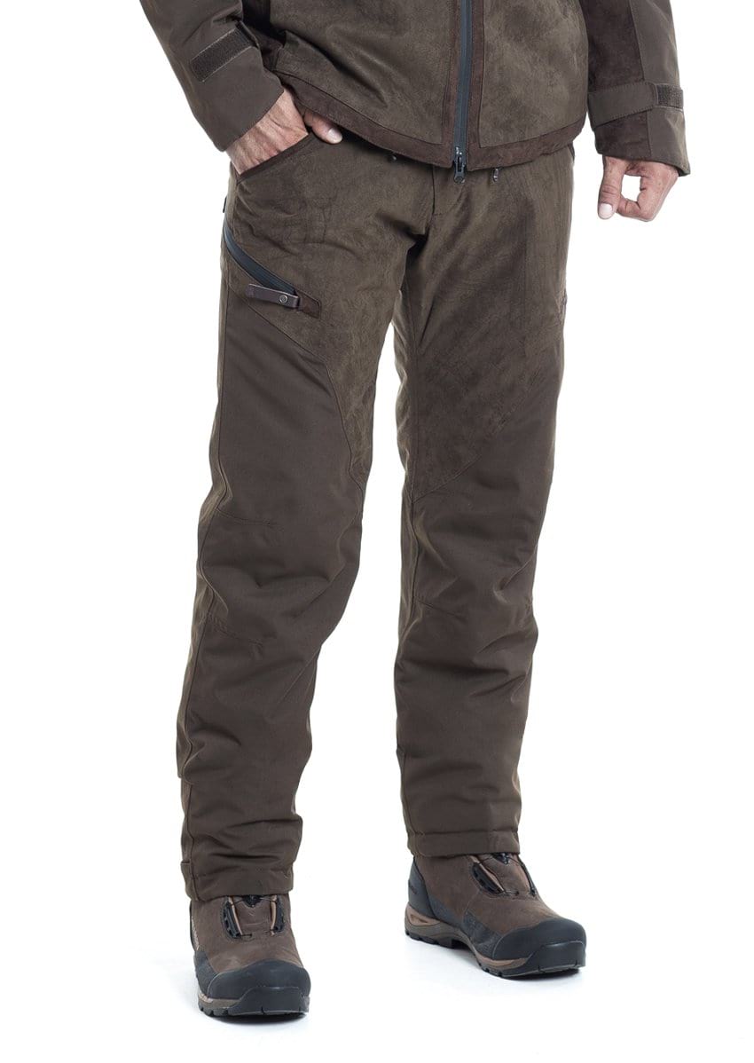 A person wears Hillman® PRE ORDER Fusion Waterproof Hunting Trousers with an ergonomic fit and a left thigh zippered pocket, along with a brown jacket and hiking boots, standing against a white background with hands at their sides.