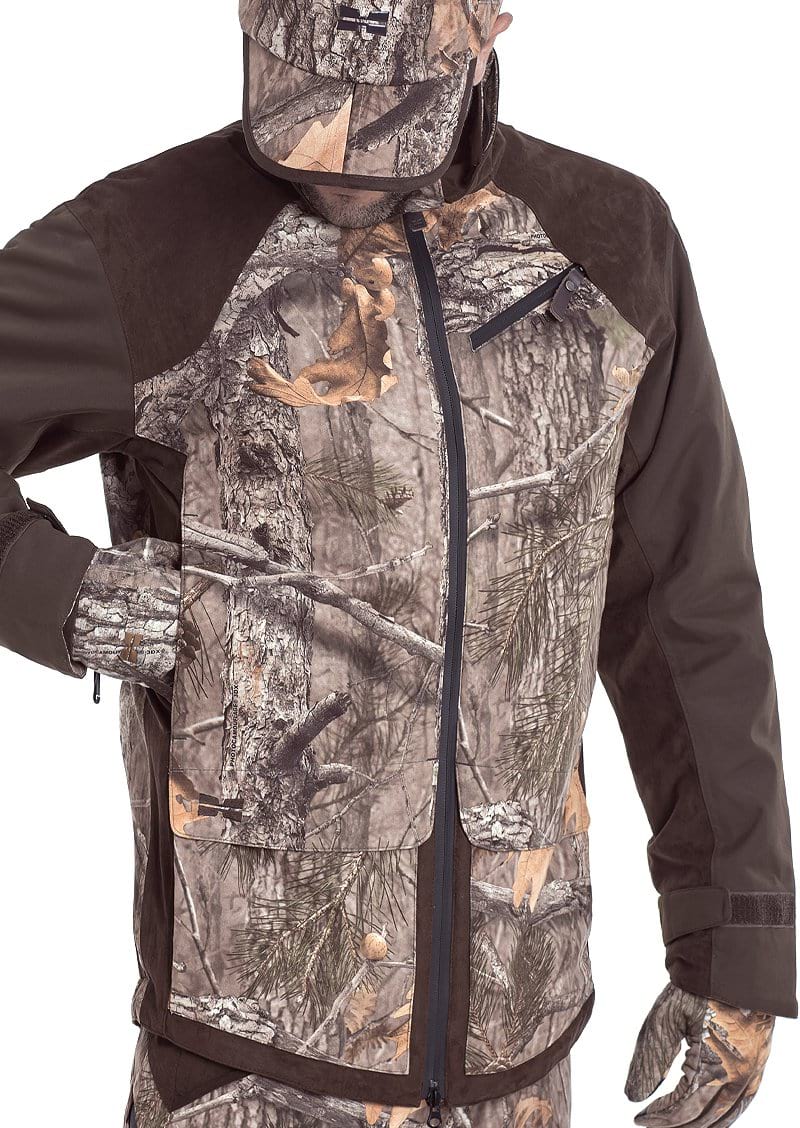Wearing the Hillman® Fusion Hunting Jacket 3DX with forest patterns, the person blends into the trees. Their face is partly covered, and one hand is tucked in the jacket.