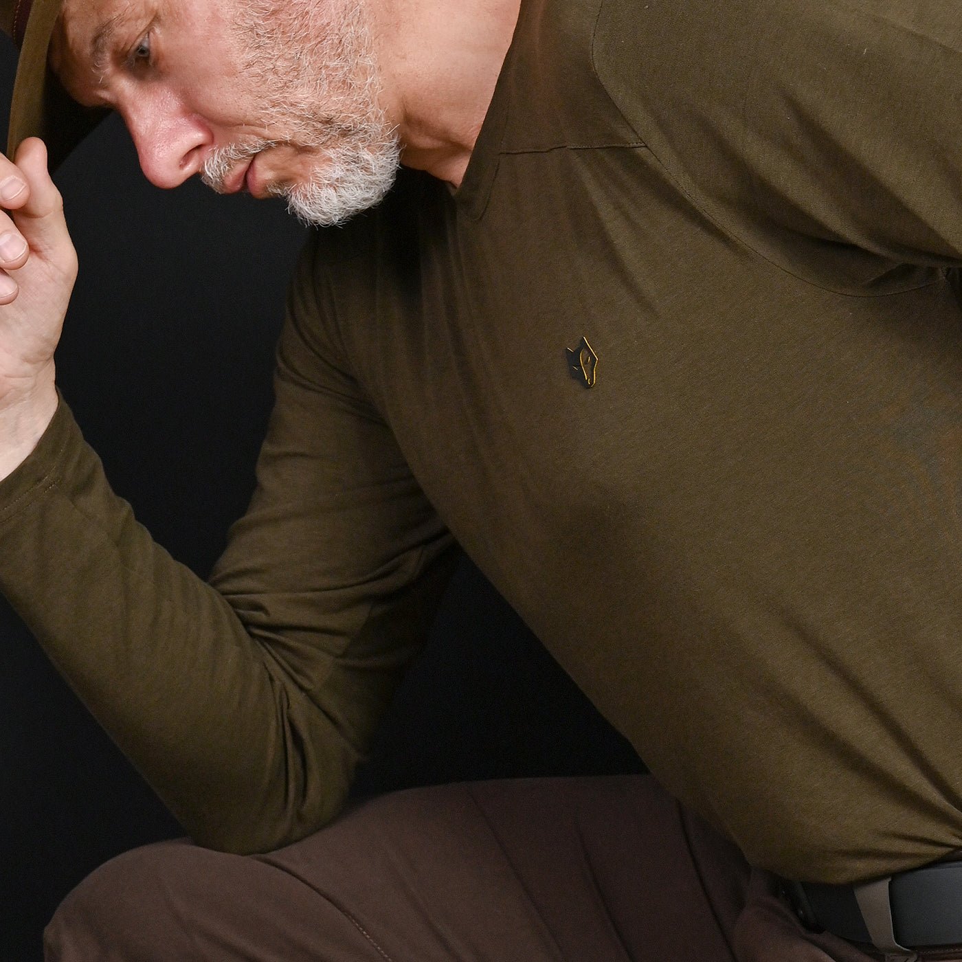 A gray-bearded man in brown pants wears the HILLMAN bamboo long sleeve hunting t-shirt—Ultralight All-Season Base Layer—and holds his hat's brim while looking down against a black background.