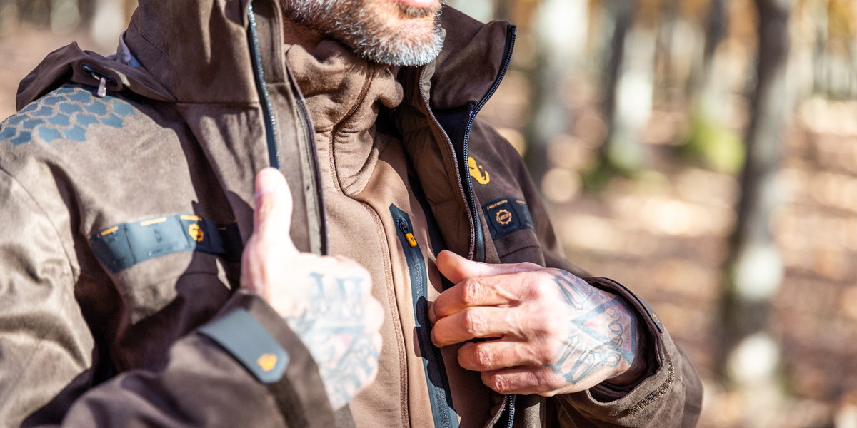 A person with tattoos on their hands adjusts the front zipper of a brown outdoor jacket in a sunlit forest, wearing a matching hoodie underneath.