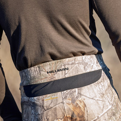 A person wears a dark long-sleeve shirt with HILLMAN GEAR waistband visible on their 5WL Ultra-Lightweight Waterproof Hunting Trousers. In the blurred outdoor backdrop, these breathable camouflage-patterned pants seem perfect for any adventure.