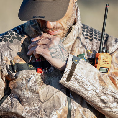 A hunter in a HILLMAN GEAR ultra-lightweight camo rain jacket holds a call device to their mouth, two-way radio and shotgun shells clipped on. Their cap-shaded face is partly visible, showing a tattooed hand.