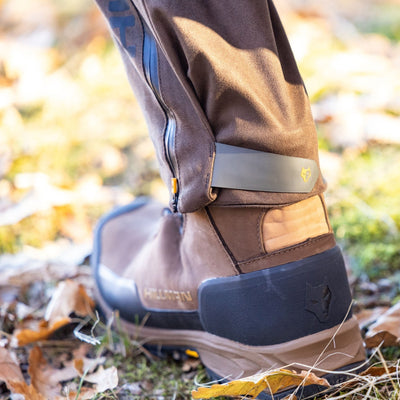Close-up of a persons lower leg in brown outdoor boots with a black sole and logo, wearing HILLMAN GEAR 6OL Matrix Insulated Hunting Trousers | Waterproof Bibs with a zipper and strapped cuff for extreme cold protection. Fallen leaves and grass are visible on the ground.