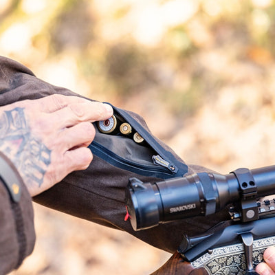 A tattooed hand unzips a pouch on the sleeve of the HILLMAN GEAR heated hunting jacket, The Warmest Heated Coat, revealing rifle cartridges inside while holding a scoped rifle. The outdoor background is blurred.