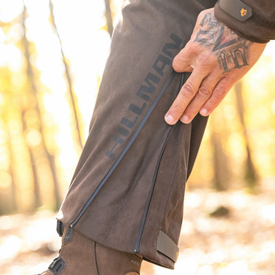 A person with tattooed fingers zips up the side of brown HILLMAN GEAR waterproof insulated hunting trousers bibs, with blurred trees and sunlight in the background.