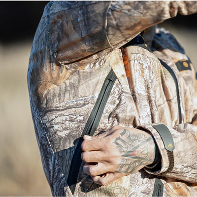 A person wearing the HILLMAN GEAR ultra-lightweight camo rain jacket unzips a sleeve pocket, their tattooed hand visible against a blurred grassland backdrop.