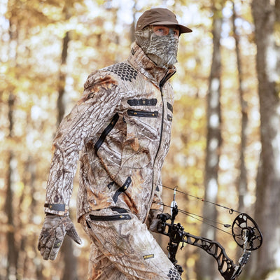 Clad in HILLMAN GEARs Matrix Insulated Hunting Trousers and face cover, a camouflaged person wields a compound bow in the forest, blending seamlessly with the autumnal backdrop—warmly protected for extreme cold.