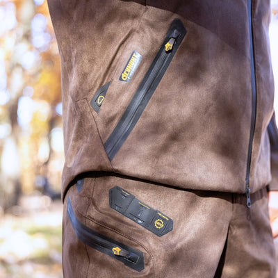 Close-up of a person outdoors wearing the HILLMAN GEAR heated hunting jacket, The Warmest Heated Coat, with matching pants. The outfit features black zippers, yellow accents, and multiple angled zippered pockets.
