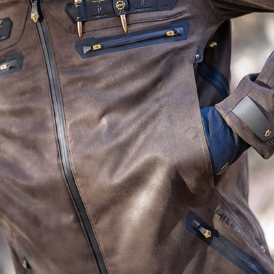 A close-up of HILLMAN GEAR’s heated hunting jacket, The Warmest Heated Coat, in brown with black zippered pockets and gold accents. Featuring detailed stitching, it's perfect for cold weather, with a gloved hand tucked into a side pocket.