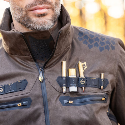 A man wears the HILLMAN GEAR heated hunting jacket, The Warmest Heated Coat, featuring chest tool holders for pens and gadgets. The blurred background glows with warm, golden tones.