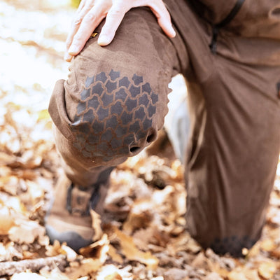 A person kneels on fallen leaves, wearing HILLMAN GEARs 6OL Matrix Insulated Hunting Trousers | Waterproof Bibs. Their hand rests on the knee, showcasing the textured patch and providing extreme cold protection in brown clothing.