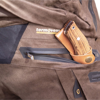 A close-up of HILLMAN GEAR insulated hunting trousers waterproof bibs, featuring a zippered pocket holding a wooden-handled knife in a leather sheath.