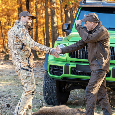 Two men in camo, wearing HILLMAN GEARs 6OL Matrix Insulated Hunting Trousers, shake hands and smile by a green SUV in an autumn forest. One doffs his cap as they stand over a large animal partially visible on the ground.