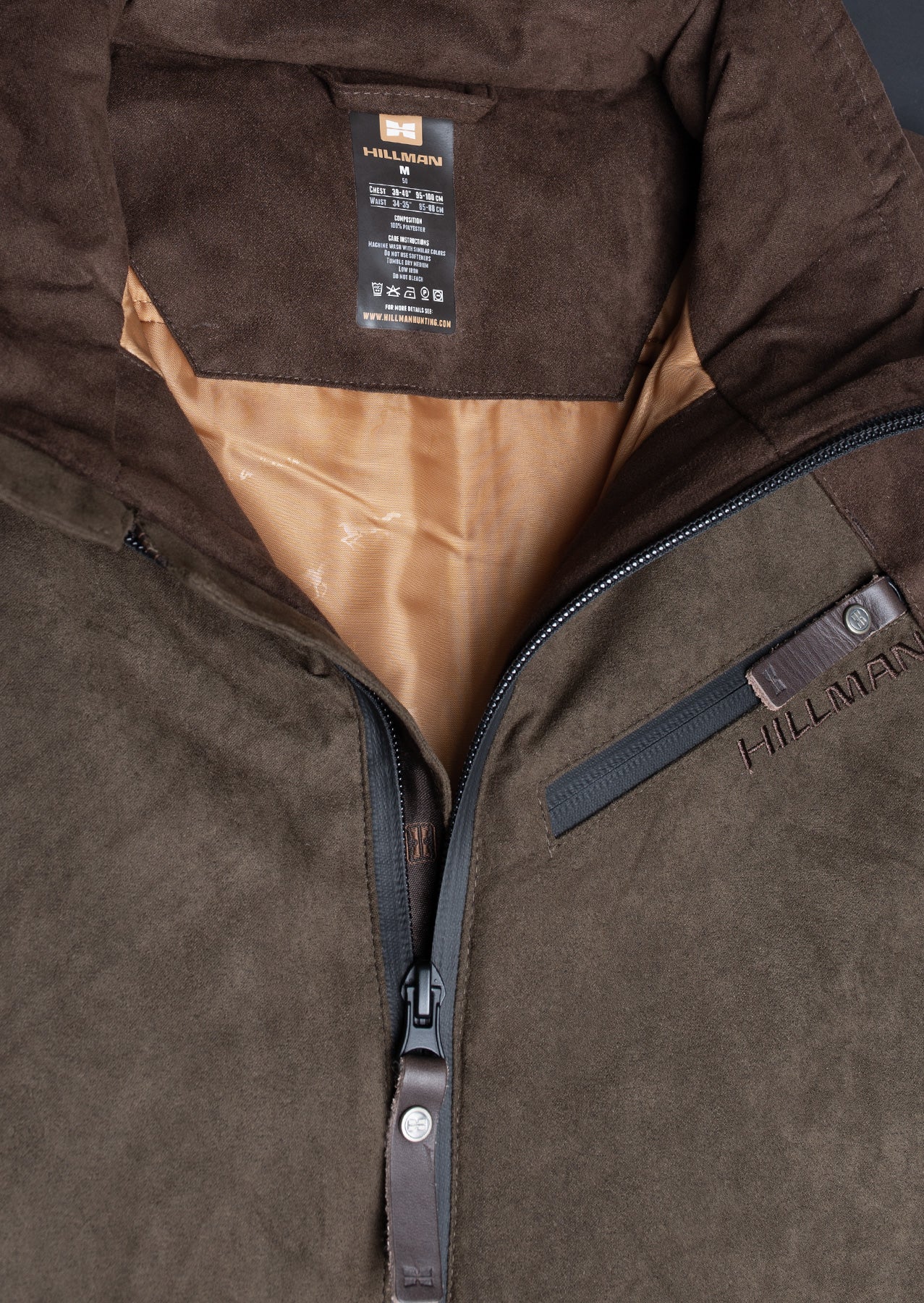 Close-up of a brown suede Hillman® waterproof hunting jacket with a partially open zipper, showing a tan inner lining and black label. The jacket features a zippered chest pocket with HILLMAN® branding.