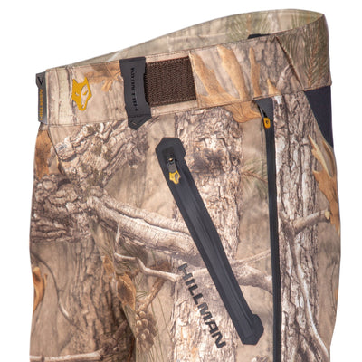 Close-up of the 5WL Ultra-Lightweight Waterproof Hunting Trousers by HILLMAN GEAR, featuring a woodland camouflage design with trees and foliage. Includes zippers and a fox symbol on the Velcro-secured waistband, along with the HILLMAN logo.