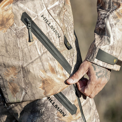 A tattooed hand unzips a pocket on a HILLMAN GEAR ultra-lightweight camo rain jacket. This waterproof camouflage hunting coat features a woodland pattern.