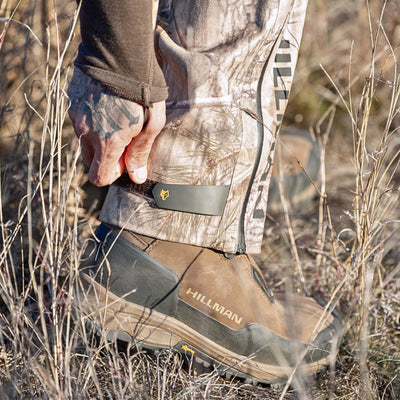 In a grassy area, someone adjusts the strap on their boot while wearing camouflage-patterned 5WL Ultra-Lightweight Waterproof Hunting Trousers by HILLMAN GEAR. The brown boots display Hillman branding, and tattooed hands emerge from under a long-sleeved shirt against a backdrop of dry grass.