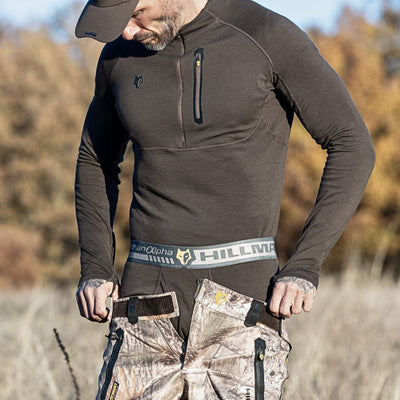 In a natural setting with trees and grass, a man adjusts his camouflage pants wearing a 2BL Merino Wool Hunting Long Johns thermal base layer shirt and cap. The waistband of the pants reads Hillman.