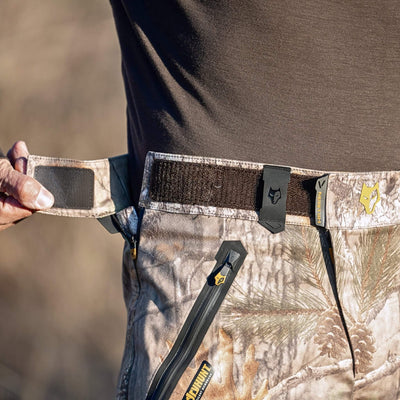 A person adjusts a camouflage-patterned belt with a Velcro strap on 5WL Ultra-Lightweight Waterproof Hunting Trousers by HILLMAN GEAR, featuring zippered pockets with yellow tabs and a stylized wolf head logo, while wearing a dark shirt.