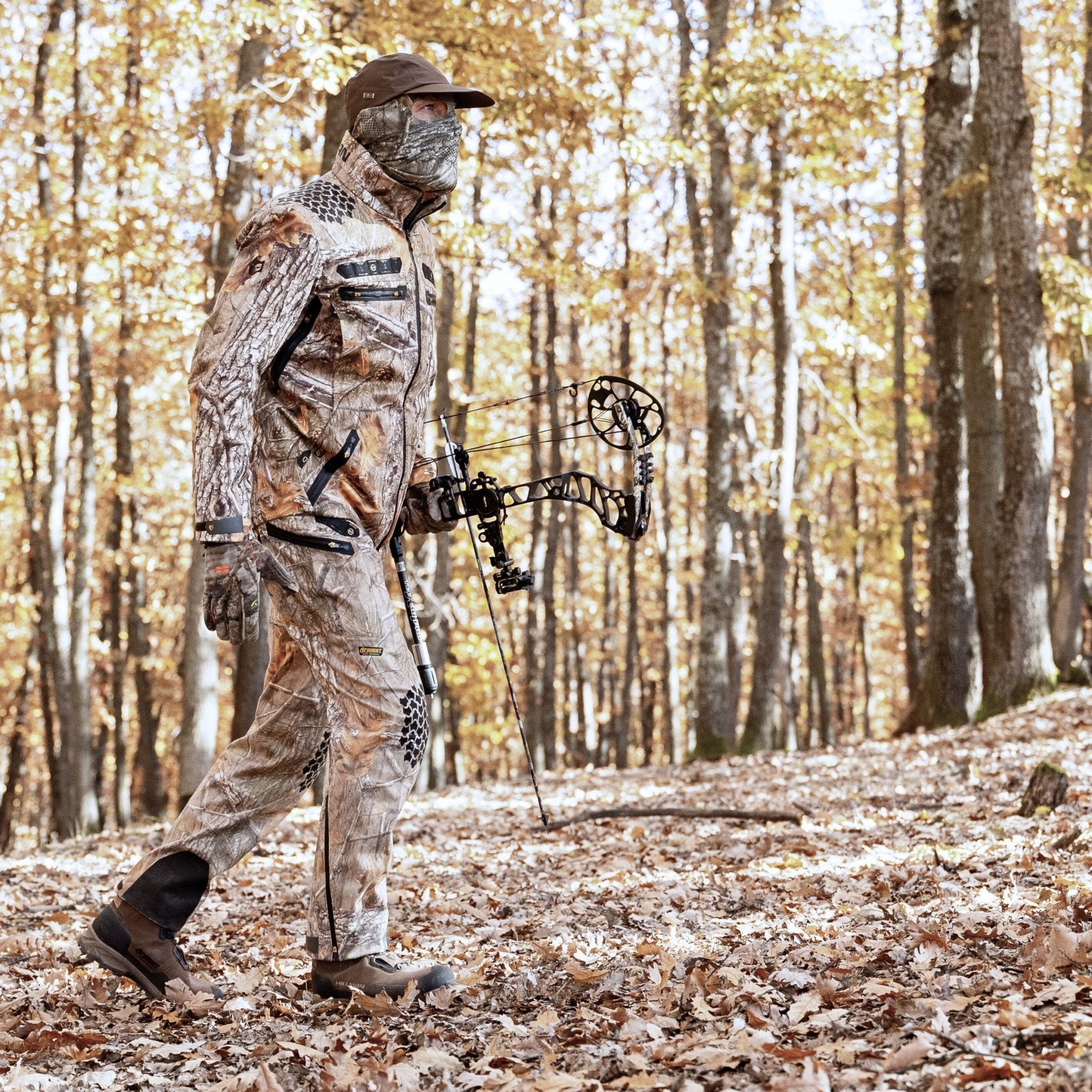 Dressed in HILLMAN GEARs 6OL Insulated Camo Hunting Trousers - Waterproof Camouflage Bibs, a person navigates an autumn forest with a compound bow. Surrounded by fallen leaves and golden foliage, theyre ready for extreme cold with a face cover and cap.