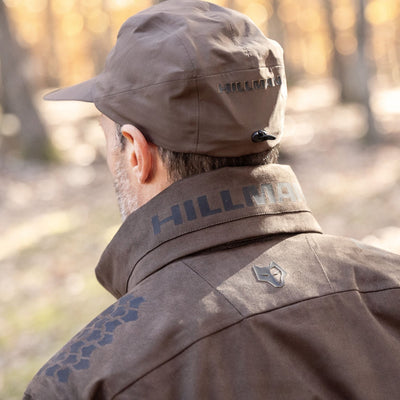 A person wearing the HILLMAN GEAR heated hunting jacket, The Warmest Heated Coat, and a matching cap stands outdoors in a sunlit, blurry forest. Viewed from behind, the jacket’s collar and logo are visible.
