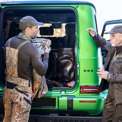 Two men in outdoor gear chat near a green SUV with its back door open. One holds HILLMAN GEARs 6OL Matrix Insulated Hunting Trousers | Waterproof Bibs, while the other gestures with a cup. Inside the vehicle are two bags and Extreme Cold Protection gear for their adventure.