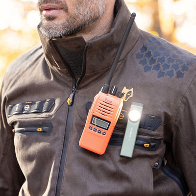 A man wears the HILLMAN GEAR heated hunting jacket, The Warmest Heated Coat—brown with hexagonal patterns—while an orange walkie-talkie and green flashlight are clipped to his chest pockets. His upper body and partially visible face are in focus.