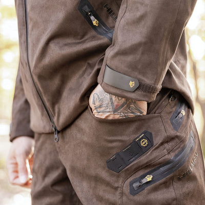 Close-up of a person wearing HILLMAN GEAR Waterproof Bibs—brown, rugged insulated hunting trousers with zipper pockets and patches. A tattooed hand rests in the pocket. Background is blurred, implying an outdoor setting.