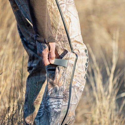 A person zips up HILLMAN GEARs 5WL Ultra-Lightweight Waterproof Hunting Trousers, adorned with nature patterns. Breathable fabric complements the rural setting of tall, dry grass in the background.
