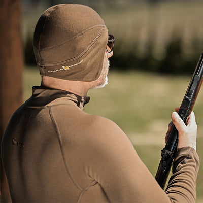 A person wearing the Hillman® Anatomic Merino Beanie [16μm] and a matching jacket holds a shotgun outdoors on a sunny day, with grass and trees blurred in the background.