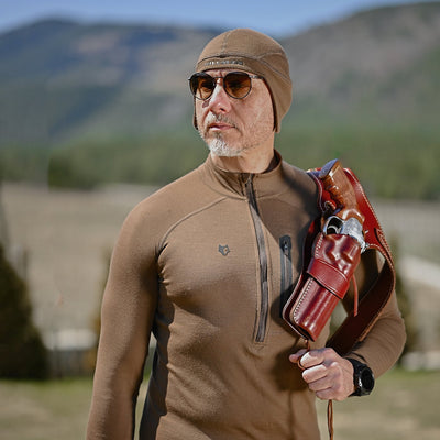 A man in a brown outfit and sunglasses wears the Hillman® Anatomic Merino Beanie [16μm], holding a leather shoulder holster with a revolver. He stands outdoors, mountains and trees blurred in the background.