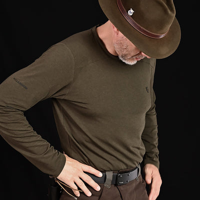 A man in a brown hat and HILLMAN bamboo long sleeve hunting t-shirt—an ultralight, moisture-controlling all-season base layer—stands against a black background, looking down with one hand on his hip, face partly hidden by the brim.